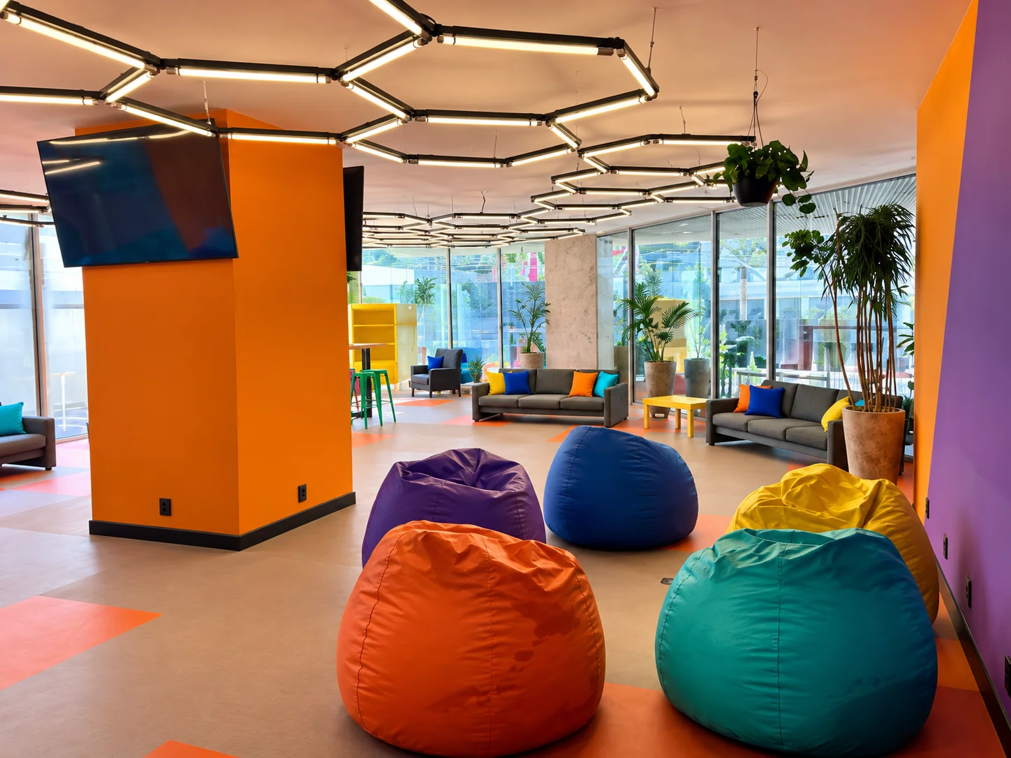 Espaço de convivência concluído, com iluminação geométrica, sofás e pufes coloridos.